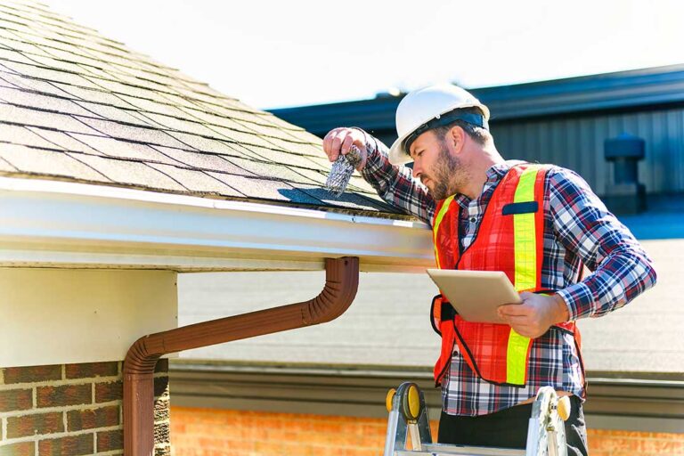 A person in safety gear using a hose and soft broom to clean moss from a residential asphalt shingle roof.