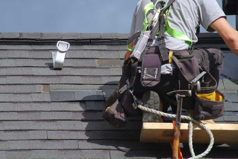 A worker standing on a sloped residential roof, examining or installing asphalt shingles under clear skies.